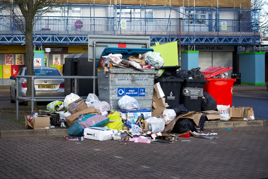 A collection of overflowing waste bins and scattered rubbish on a paved sidewalk in front of a commercial building, with a silver car parked nearby. The waste containers consist of a large blue bin la