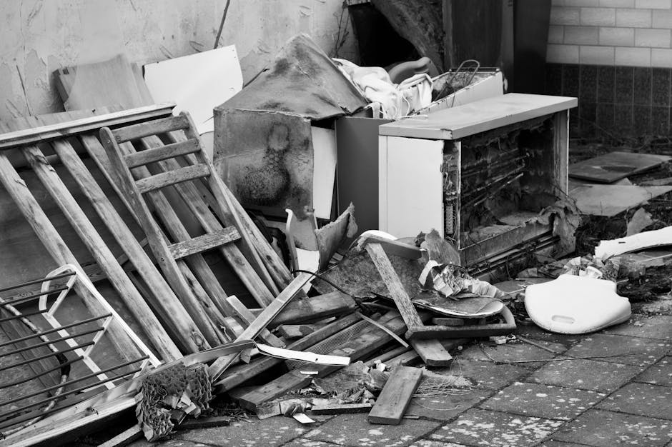 A pile of discarded household items and debris on a paved surface outside a building, including a dismantled wooden ladder leaning against the wall, broken wooden planks, and various pieces of damaged