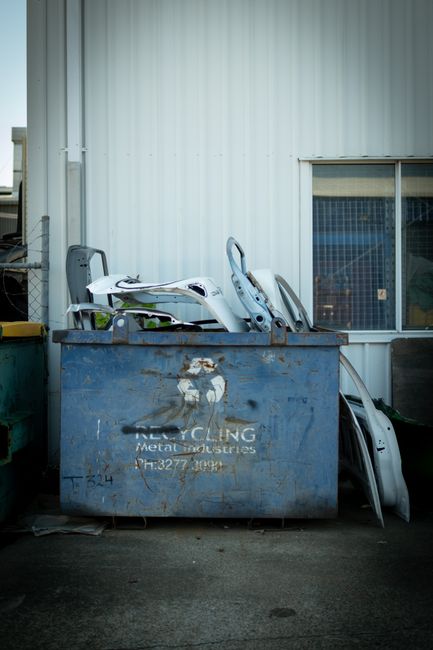 A collection of used black rubber tires is stacked inside two large, industrial blue metal containers positioned outdoors in front of a white corrugated metal building with moss and dirt accumulation 