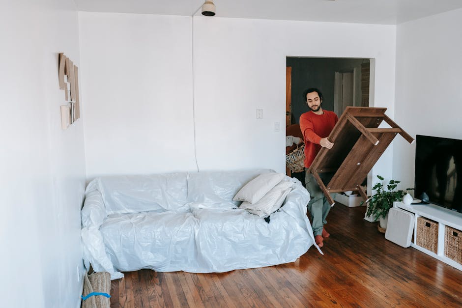 A man with a beard and casual clothing is lifting a small wooden table with a natural wood finish, which is being removed from a living room. The room features white-painted walls and a polished woode